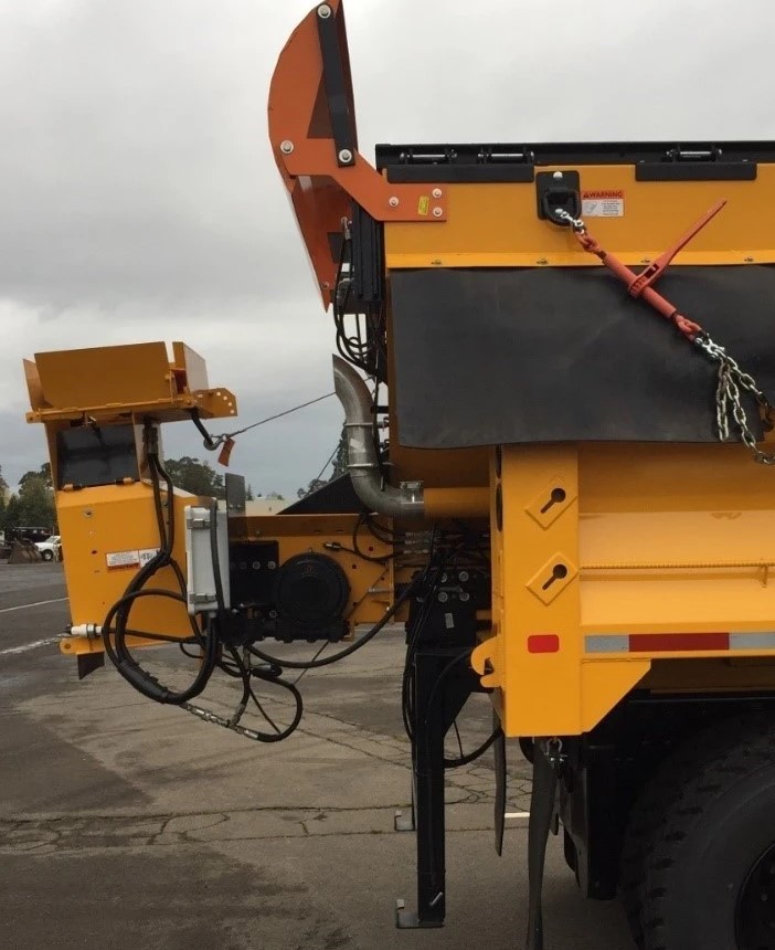 An image of the side of a yellow PennDOT plow truck with an orange slightly curved metal barrier mounted to the top protecting the truck from a buildup of snow on the rear of the vehicle